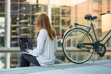 Fototapeta na wymiar a young charming woman sitting near the river somewhere in the city and working with a laptop; her bicycle is standing next to her