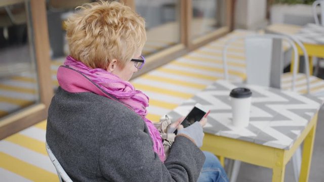 Adult blonde woman browsing smartphone and having drink from paper cup in outside cafe.