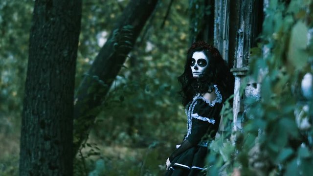 The Mexican Day of the Dead. The young woman with spooky sceleton make-up for Halloween dressed in black clothes leaning to wooden wall and looking at camera. 4K