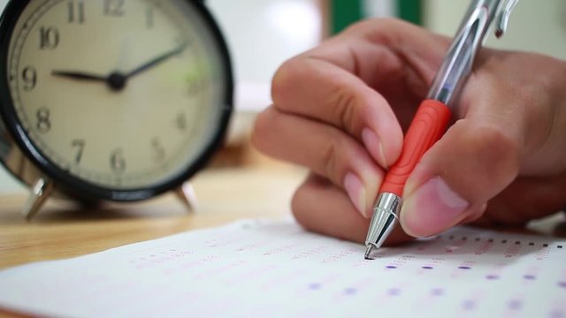 Asian students hands holding pen taking fill drawing selected of choice on Exams paper sheet or test paper on wood desk in school, college with retro clock. Education learning concept.