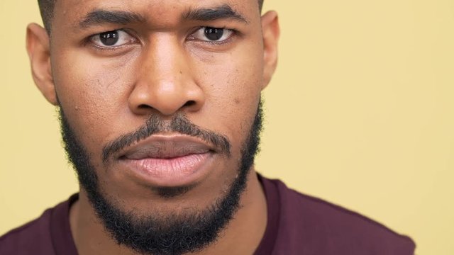 extreme closeup portrait of confident african american guy with brown eyes wearing sporty clothes looking strictly at camera blinking standing over yellow background slomo. Concept of emotions