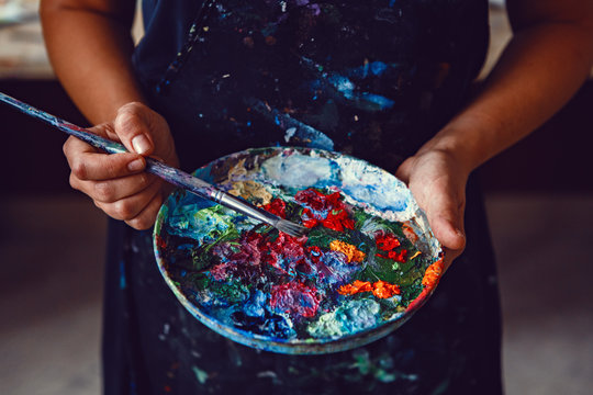 Hands of female artist holding messy dirty palette  with different paints and paintbrush in art studio. Lifestyle and hobby concept
