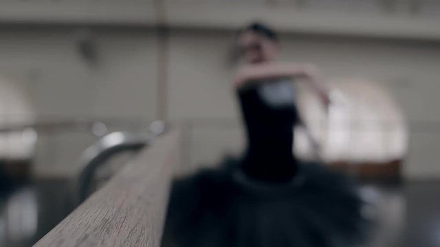 Ballerina in black tutu stretches on barre in ballet gym. Woman standing near bar, preparing for practice. Blurred video. Slow motion.