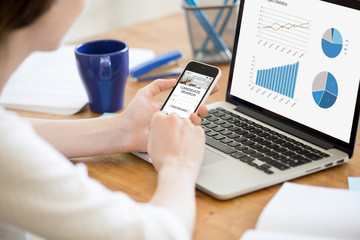 Business woman sitting in front of laptop with project statistics data, using smartphone phone...