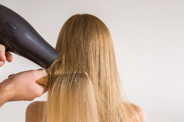 Naklejka premium Beautician's hands using hair dryer and comb on woman's blonde hair on the gray background. Cares about a healthy and clean hair. Beauty salon concept.