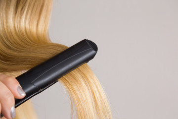 Woman's hand using straightener on her blonde hair on the gray background. Cares about a smooth, beautiful hair. Beauty salon concept. © fotoduets
