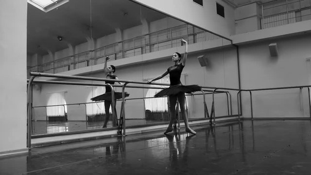 Ballerina in black tutu and pointe stretches on barre in ballet gym. Woman standing near bar and mirror, preparing for perfomance. Slow motion. Black and white tonned footage