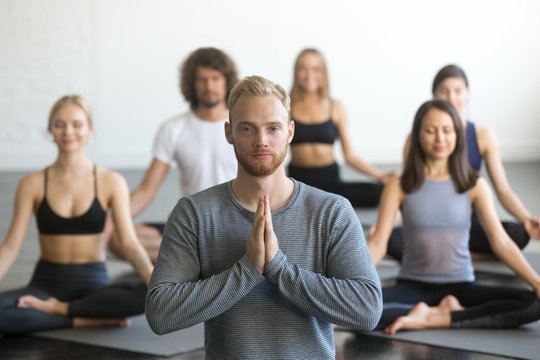Group of young sporty people practicing yoga lesson with instructor, sitting in Sukhasana exercise, Easy Seat pose, friends working out in club, focus on male student, making mudra gesture, studio