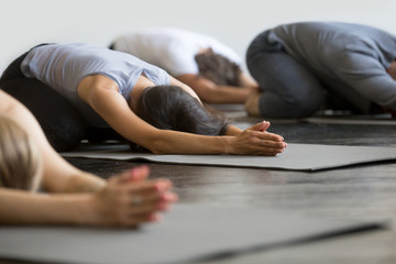 Group of young sporty people practicing yoga lesson with instructor, sitting in Balasana exercise, Child pose, friends working out in club, indoor close up image, studio. Wellbeing, wellness concept - obrazy, fototapety, plakaty