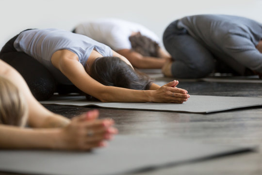 Group of young sporty people practicing yoga lesson with instructor, sitting in Balasana exercise, Child pose, friends working out in club, indoor close up image, studio. Wellbeing, wellness concept