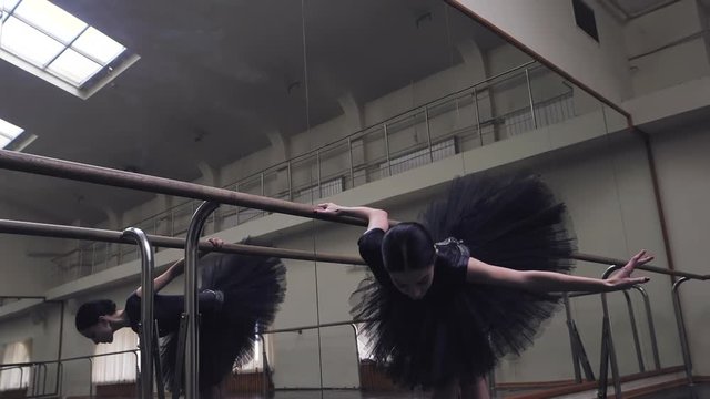 Ballerina in black tutu and pointe stretches on barre in ballet gym. Woman standing near bar and mirror, preparing for perfomance. Slow motion.