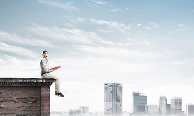 Man on roof edge reading book and cityscape at background