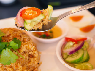 Close up spoon scooping Thai local traditional style stir fried rice with egg and fermented pork and nuts with coconut milk curry paste, “Sa Tae”, with chili, cucumber vinegar, in white ceramic plate