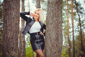 A young sexy woman in a forest is dressed in a leather jacket and a skirt demonstrates herself and clothes in a forest against a background of pines