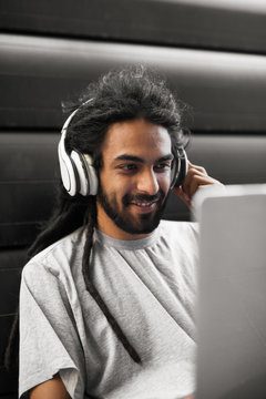 Casual indian hipster sitting on the street. Holding headphone looking at laptop smiling.