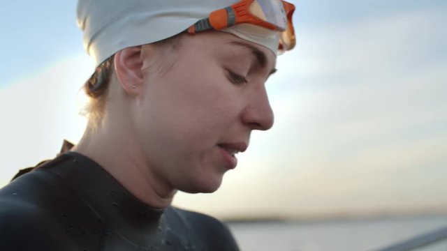 Close up of face of female swimmer in cap and goggles sitting on boat and wincing while relieving foot cramp after practice in open water