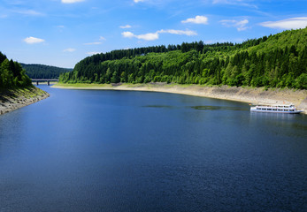 Fototapeta premium Landscape with a lake surrounded by green fresh fir trees on a beautiful sunny summer day in the harz national park. White ship transporting tourists crossing by. 