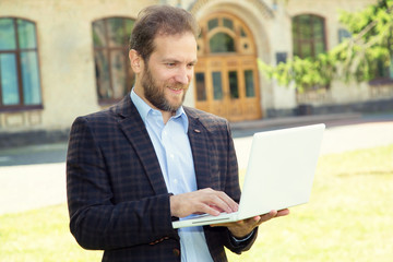 Business expert. Confident handsome man with beard in shirt hold laptop and smiling while standing against university background.