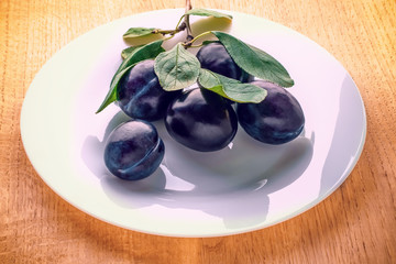 Ripe plums in a white plate with green leaves on a branch on a wooden background.