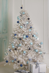 A decorated Christmas tree stands near a bright wall. Artificial white spruce with blue balls and garlands.