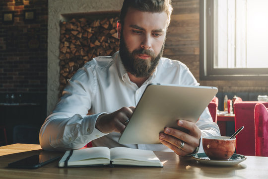 Young bearded businessman sits in office at table,uses tablet computer. On desk is notebook,smartphone, cup of tea.Man working, studying.Online education,marketing, training. E-learning, e-commerce.
