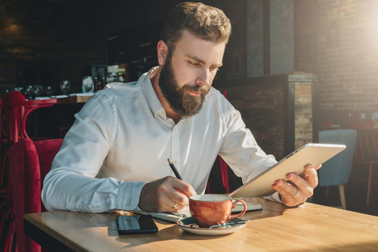 Young bearded businessman sits in cafe, home at table, uses tablet computer and writes in notebook. Man is working, student is studying. Online education, marketing, training. E-learning, e-commerce.