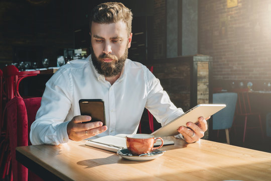 Young bearded businessman,dressed in white shirt, sits in cafe at table, holds digital tablet, uses smartphone.There is notebook on table,cup of coffee.Online education,marketing,training.E-learning.