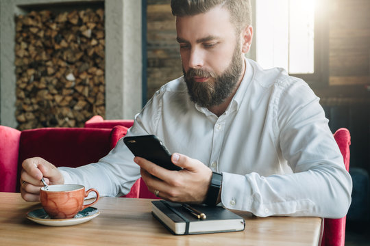 Young bearded businessman, dressed in white shirt, sits in cafe, office at table,reads information on smartphone screen, drinks coffee.On desk is notebook, pen.Online marketing, education, e-learning.