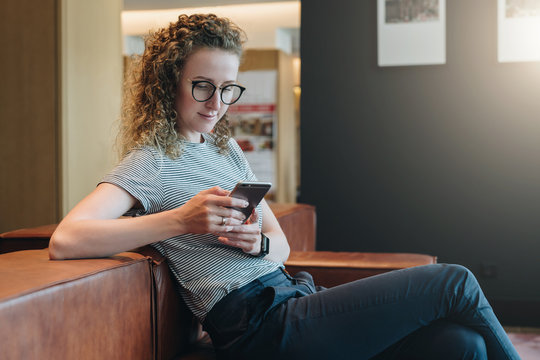 Young smiling woman with curly hair in eyeglasses sits in waiting room on couch and uses smartphone. Hipster girl working, studying, checking email.Online marketing, education. E-learning, e-commerce.