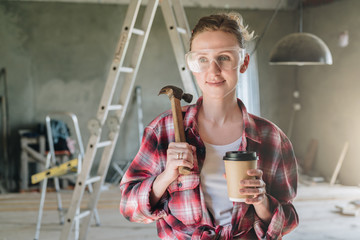 Front view. Portrait of happy young female construction worker, carpenter, repairman standing in workshop wearing safety glasses, holding hammer and cup of coffee in his hand.In background stepladder.