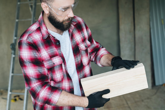Young bearded businessman, builder, repairman, carpenter, architect, designer dressed in plaid shirt, glasses and gloves, stands in workshop, holds a wooden cast and looks at it.