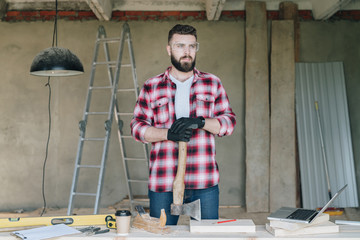 Young bearded businessman, builder, repairman, carpenter, architect, designer dressed in plaid shirt, goggles and gloves, stands in workshop and holds ax, hatchet in his hand. Handyman working.