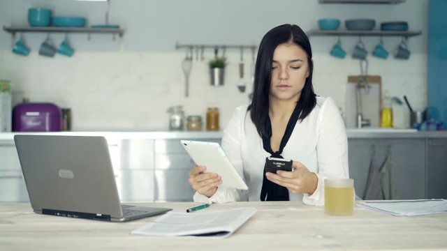 Young businesswoman comparing data on tablet and smartphone by table at home
