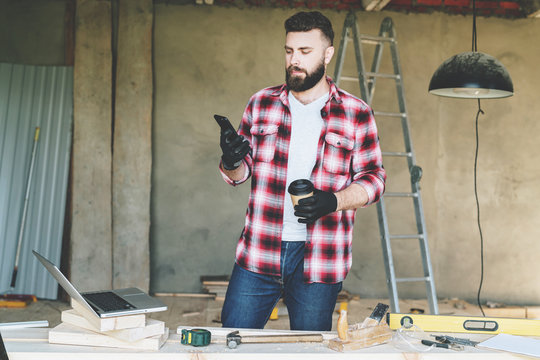 Young bearded businessman, builder, repairman, carpenter, architect, designer dressed in plaid shirt, goggles and gloves, stands in workshop, using smartphone and holds cup of coffee in his hand.