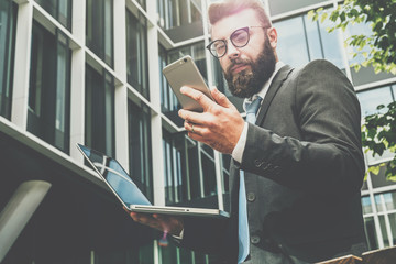 Young bearded businessman stands against modern building, uses smartphone and holds a laptop in his hand. Man works remotely. Distance job. Online marketing, education,training,e-commerce,e-learning.