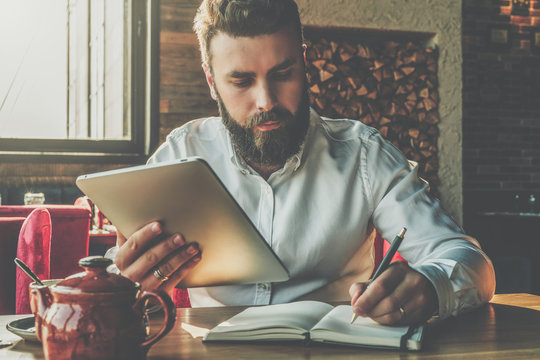Young bearded businessman sits in cafe at table, holding tablet computer and writes in notebook. Man is working, studying.Online education, marketing, training.E-learning,e-commerce. Instagram filter.