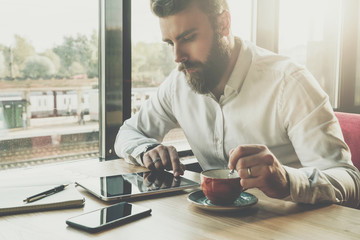 Young bearded businessman sits in office at table,uses tablet computer.On desk is notebook,smartphone.Man working,studying.Online education,marketing,training.E-learning, e-commerce. Instagram filter.