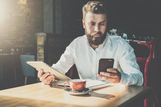 Young serious bearded businessman sits in cafe at table, holds digital tablet, uses smartphone.There is notebook on table,cup of coffee.Online education,marketing,training.E-learning.Instagram filter.