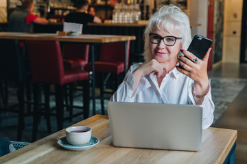 Businesswoman in glasses is sitting at table in front of laptop and holding smartphone.Education for adults. Freelancer works. Retired woman is chatting, blogging, checking email.Social media, network