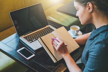 Side view. Young businesswoman sitting at table in cafe in front of laptop, computer and making notes in notebook, diary. Girl working, student studying. Online marketing, education, e-learning.