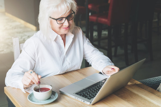 Smiling businesswoman in glasses sitting at table, working on laptop and drinking tea. Education for adults. Pensioner freelancer works. The woman is chatting, blogging, checking email. E-marketing.