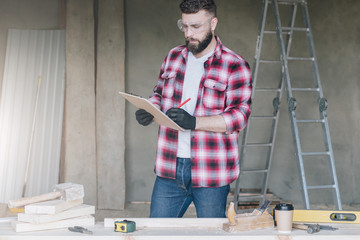 Hipster bearded man is carpenter, builder, designer stands in workshop, holds clipboard and takes notes. On desk is construction tools, in background stepladder. Repair, construction, carpentry.