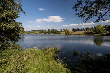 pond between woods and fields with a small cottage on the bank in summer