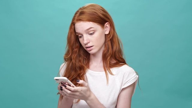 Displeased ginger woman in t-shirt writing message on smartphone over turquoise background