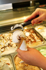 Female hand with scoop takes ice cream from the fridge and serving in waffle cone. Woman taking scoop of tasty ice cream. Fridge with ice cream. Woman works in ice cream shop. Pastry shop. Desserts.