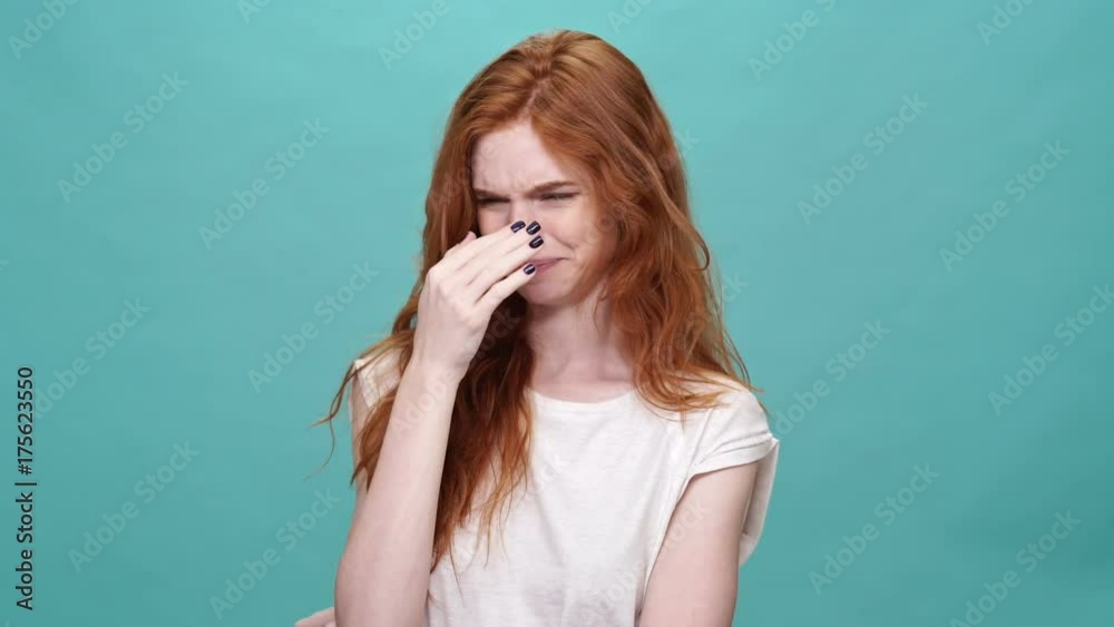 Wall mural Pretty ginger woman in t-shirt covering her nose because of unpleasant odor over turquoise background