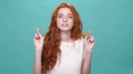 Young ginger woman in t-shirt praying with crossed fingers and upset after thatover turquoise background
