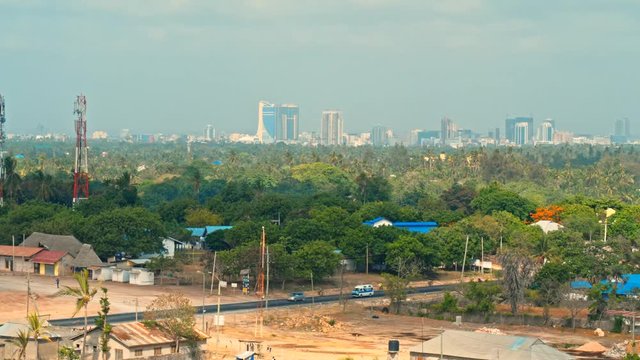 Cinematic aerial of port city Dar es Salaam downtown skyline with skyscraper buildings, lush, green vegetation, palm trees, minor vehicle traffic near Indian ocean beach, Tanzania, Africa