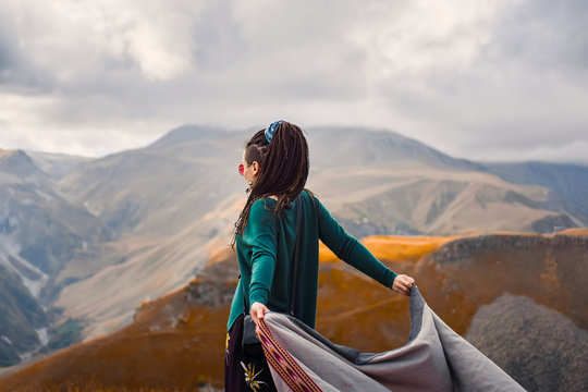 portrait of a young beautiful hipster girl in sunglasses with hair dreadlock in a boho gypsy hippie style clothes on a background of cloudy sky and autumn mountains of Georgia dancing