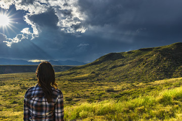 woman looks out toward stunning views of sun bursting through clouds over a mountain range - 175626793